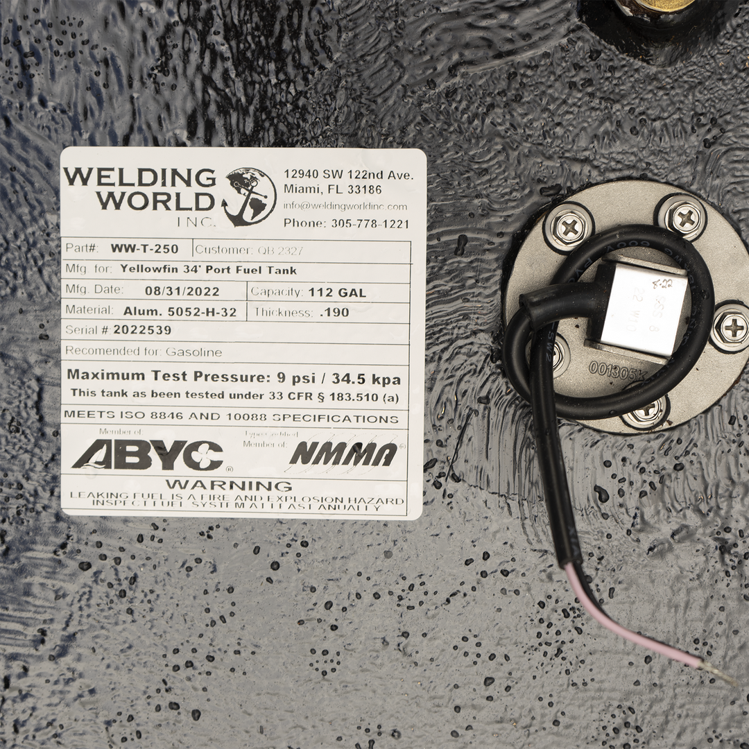 A close-up view of a product label from 'Welding World Inc.' attached to a textured black surface. The label provides detailed specifications for a 'Yellowfin 34' Port Fuel Tank,' including Part # WW-T-250, Mfg. Date 08/31/2022, Capacity 112 GAL, Material Alum. 5052-H-32, Thickness .190, and Serial # 2022539. It also states the tank is recommended for Gasoline, has a Maximum Test Pressure of 9 psi / 34.5 kpa, and meets ISO 8846 and 10088 specifications. Logos for ABYC and NMMA are present