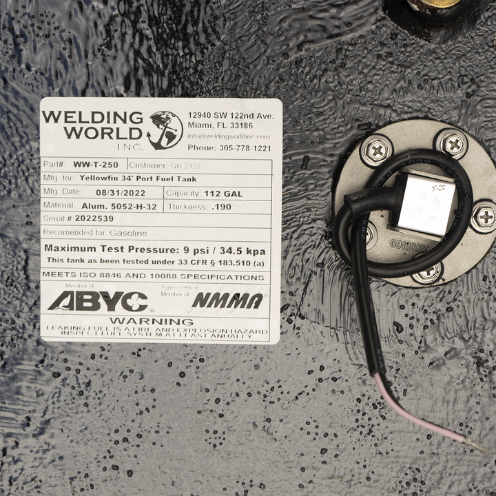 A close-up view of a product label from 'Welding World Inc.' attached to a textured black surface. The label provides detailed specifications for a 'Yellowfin 34' Port Fuel Tank,' including Part # WW-T-250, Mfg. Date 08/31/2022, Capacity 112 GAL, Material Alum. 5052-H-32, Thickness .190, and Serial # 2022539. It also states the tank is recommended for Gasoline, has a Maximum Test Pressure of 9 psi / 34.5 kpa, and meets ISO 8846 and 10088 specifications. Logos for ABYC and NMMA are present